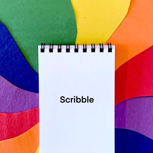May include: A white spiral notebook with the word "Scribble" printed on the front. The notebook is placed on a colorful background of rainbow-colored paper.