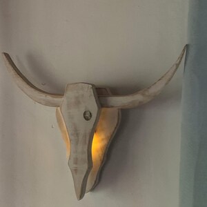 Può includere: Decorazione da parete a forma di teschio di toro in legno, con lunghe corna ricurve. Il teschio è dipinto di bianco con una finitura invecchiata, e una luce calda brilla dall'interno della cavità del teschio. Questo oggetto decorativo aggiunge un tocco di fascino western.
