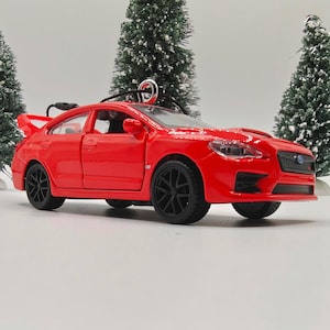 May include: A vibrant red toy car with black wheels and a rear spoiler. The car is a miniature model, possibly an ornament, with a silver loop for hanging. Two small, artificial Christmas trees are in the background.