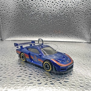 May include: A blue toy race car with gold and red accents, a rear spoiler, and the number 75 on the side. The car has a silver string for hanging, suggesting it's an ornament. The wheels are black with gold rims.