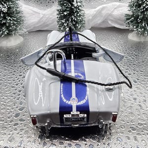 K&ouml;nnte beinhalten: Ein wei&szlig;es und blaues Modellauto mit einem schwarzen Seil, das an der Oberseite befestigt ist. Das Auto hat einen blauen Streifen in der Mitte und das Wort "COBRA" auf der R&uuml;ckseite.