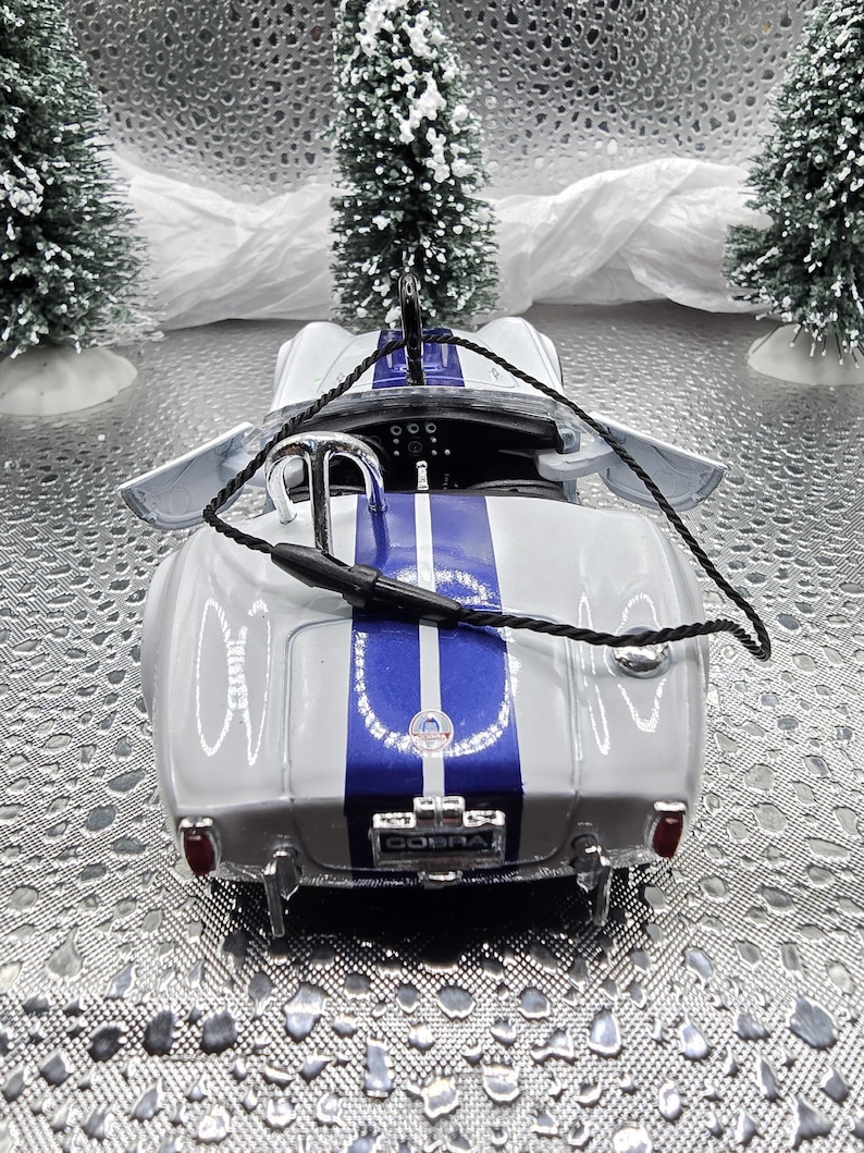 K&ouml;nnte beinhalten: Ein wei&szlig;es und blaues Modellauto mit einem schwarzen Seil, das an der Oberseite befestigt ist. Das Auto hat einen blauen Streifen in der Mitte und das Wort "COBRA" auf der R&uuml;ckseite.