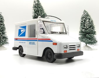 Puede incluir: Un camión de correo de juguete blanco del United States Postal Service con el logo del águila azul, rayas rojas y azules, y el texto www.usps.com y United States Postal Service. El vehículo tiene ruedas negras, un parachoques negro y un parabrisas transparente.
