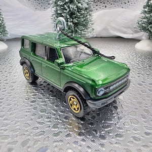 May include: A green toy SUV with gold-colored wheels and black accents. The vehicle has a shiny finish and a black rope attached to the top for hanging. The SUV is a Christmas ornament.