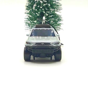May include: A silver toy pickup truck ornament featuring a black front bumper, a Toyota emblem on the grille, and dark-tinted windows. The vehicle is positioned in front of a small, snow-dusted artificial pine tree with a silver hanging loop.