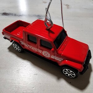 Peut inclure: Camion jouet rouge avec une étiquette blanche "EAST LAKE FIRE DEPARTMENT FIRE & RESCUE" et une bande blanche sur le côté. Le camion a un crochet argenté pour l'accrocher.