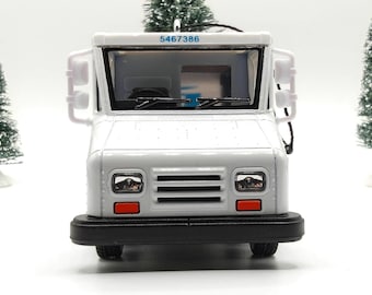 May include: A white miniature delivery truck ornament featuring a black front bumper, two red rectangular lights, and the number 5467386 printed in blue above the windshield. The vehicle has side mirrors and a visible steering wheel inside.