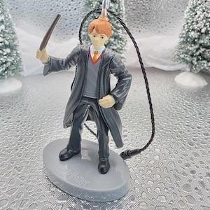May include: A figurine of a character from the Harry Potter series, wearing a black robe and holding a wand. The figure has red hair and is standing on a gray base. A black string is attached for hanging. The base has the words "Harry Potter" printed on it.