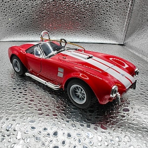 K&ouml;nnte beinhalten: Rotes und wei&szlig;es Spielzeugautomodell eines klassischen Sportwagens mit einem wei&szlig;en Streifen &uuml;ber die Motorhaube. Das Auto hat eine schwarze Innenausstattung und silberne R&auml;der. Das Auto hat die Nummer "427" an der Seite.