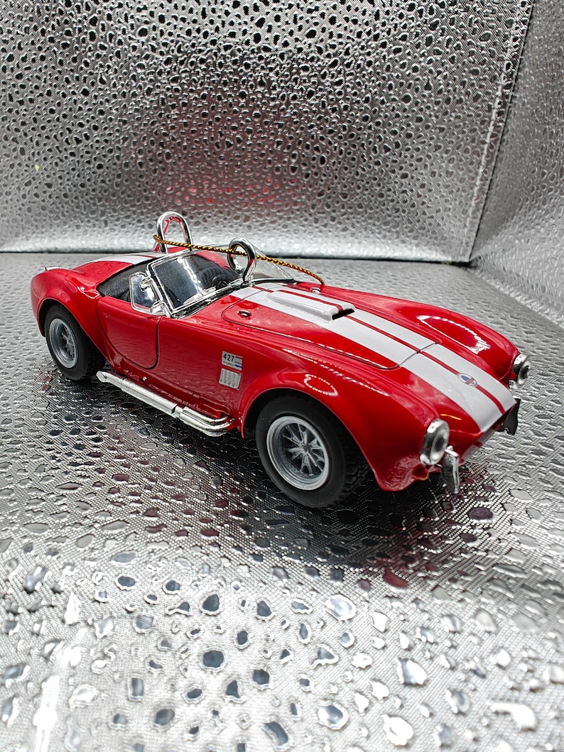 K&ouml;nnte beinhalten: Rotes und wei&szlig;es Spielzeugautomodell eines klassischen Sportwagens mit einem wei&szlig;en Streifen &uuml;ber die Motorhaube. Das Auto hat eine schwarze Innenausstattung und silberne R&auml;der. Das Auto hat die Nummer "427" an der Seite.