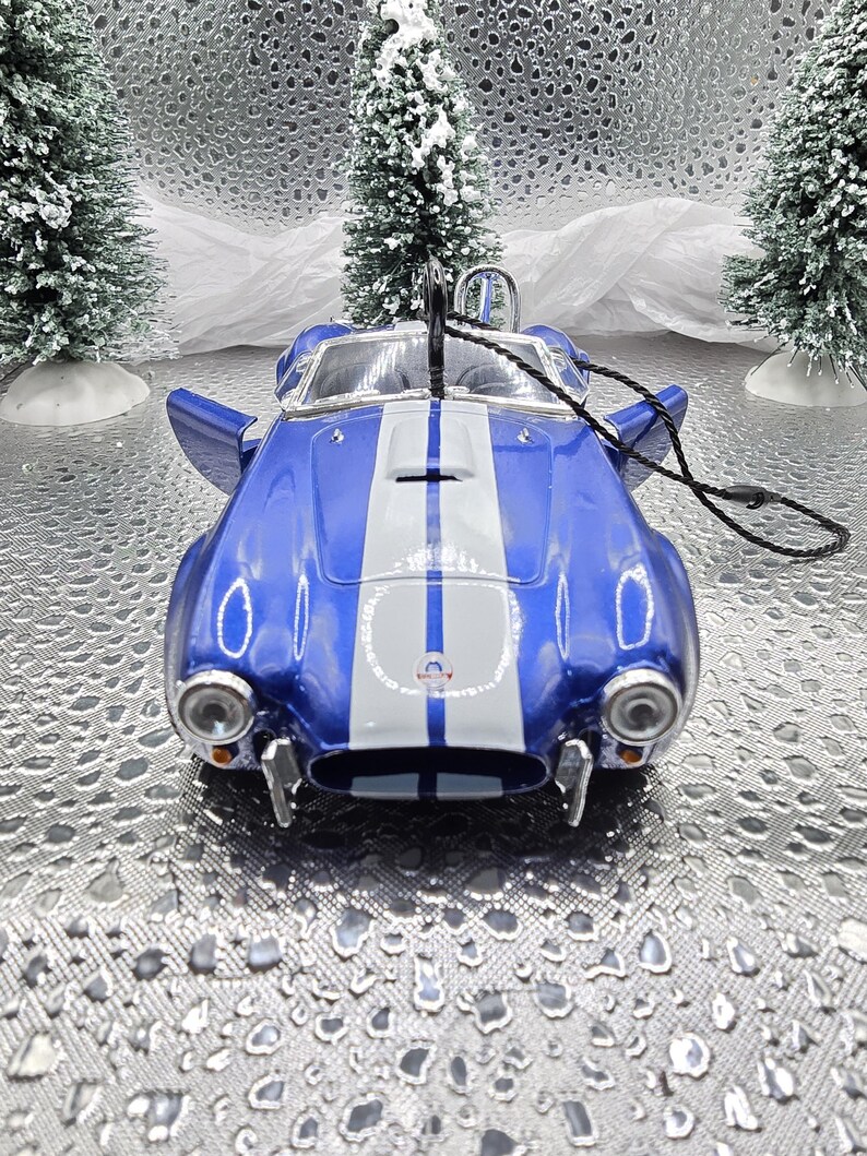 K&ouml;nnte beinhalten: Blau-wei&szlig;es Modellfahrzeug-Ornament mit einem wei&szlig;en Streifen in der Mitte der Motorhaube. Das Auto hat ein schwarzes Band, das an der Oberseite befestigt ist.