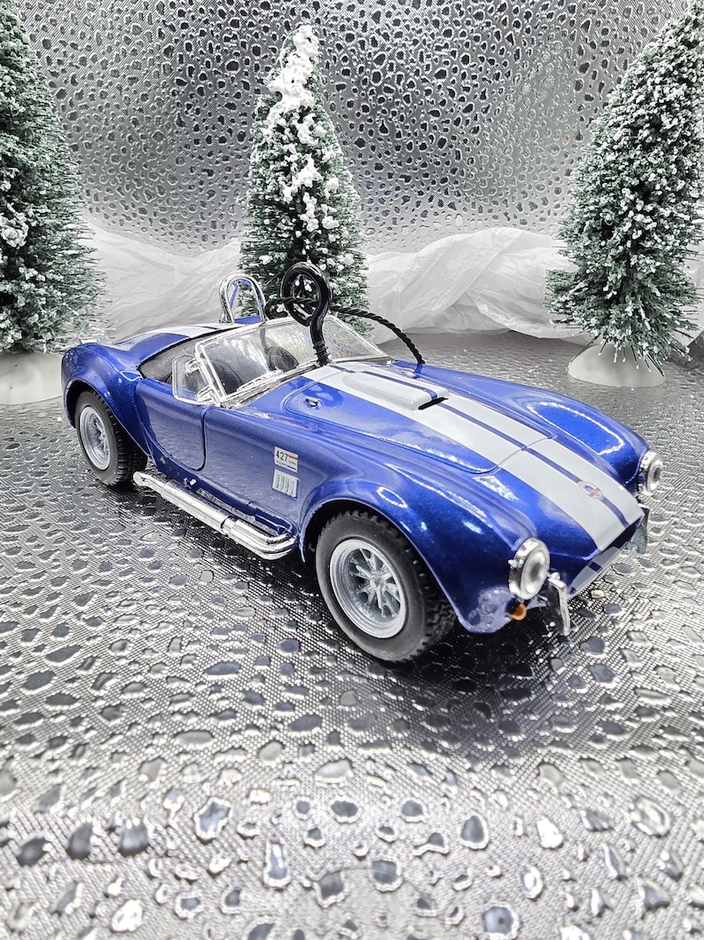 K&ouml;nnte beinhalten: Blau-wei&szlig;es Modellwagen-Ornament eines klassischen Sportwagens mit einem wei&szlig;en Streifen &uuml;ber der Motorhaube. Das Auto hat einen schwarzen Innenraum und eine schwarze Drahtschlaufe zum Aufh&auml;ngen.