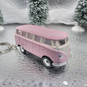 May include: A pink and cream-colored miniature Volkswagen bus keychain. The toy bus features a classic design with windows and wheels. The keychain is made of metal and has a silver keyring attached.