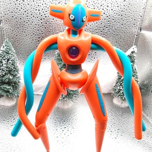 May include: Orange and teal Deoxys figure with a purple center. The figure has a unique design with a head, torso, arms, and legs. The arms are teal and orange, and the legs are orange with teal accents. The figure is a collectible item.