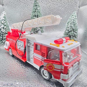 May include: A red and silver toy fire truck with a white ladder extended upwards. The toy has the words "FIRE DEPT. 119" printed on the side. The truck has a red light bar on top and is set against a silver background.