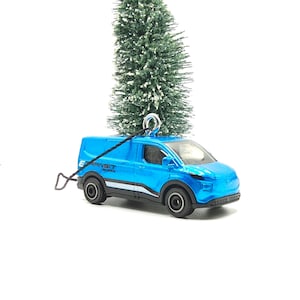 May include: A blue toy van with the text "E-Transit" on the side, designed as an ornament. The van has black wheels and a silver hook for hanging. Two small Christmas trees are in the background.