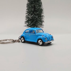 May include: A light blue toy car keyring. The miniature car has a classic design with rounded features and a silver key ring. The car is set against a white background with small decorative trees.