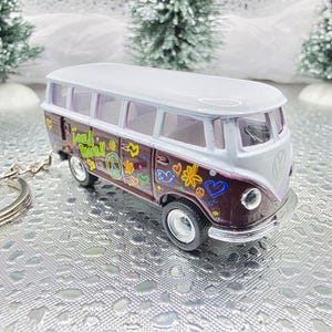 Peut inclure: Un mini-bus Volkswagen bicolore, marron et blanc, sur le thème de la paix et de l'amour. Le bus présente des décalcomanies florales et des signes de paix colorés. Le texte "Peace & Love" est visible. C'est un porte-clés.