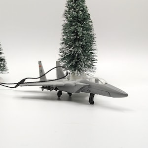 May include: A gray fighter jet model ornament featuring a clear cockpit canopy, landing gear, and a black hanging cord attached to the tail section. The aircraft has visible markings on the vertical stabilizers and wings, designed for display.