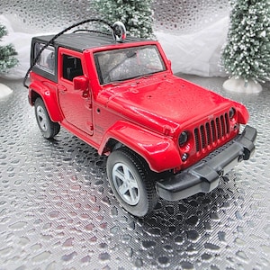 May include: A red Jeep Wrangler toy ornament with a black top and silver wheels. The miniature SUV has a black string for hanging and is set against a winter-themed backdrop with small Christmas trees.