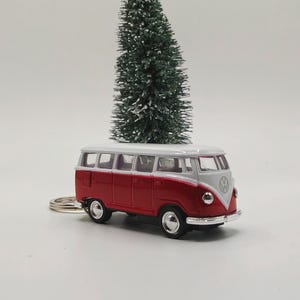 May include: A red and white miniature Volkswagen bus keychain. The toy bus has black wheels and a silver-colored bumper. The keychain is set against a white background with small Christmas trees.
