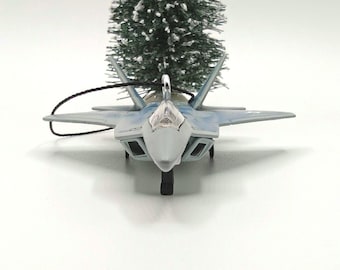 May include: A gray and blue fighter jet ornament featuring a clear cockpit canopy, angular wings, and a black hanging string attached to a silver metal loop. The model aircraft is positioned in front of a small, snow-dusted artificial pine tree.