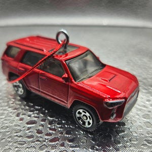 May include: A small, red SUV toy with black wheels and a silver loop for hanging. The miniature vehicle has a detailed design, including a sunroof and a hood vent. A red string is attached to the loop.