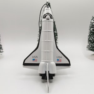 May include: A white space shuttle ornament featuring black trim, a cockpit window, and the text USA with a small American flag graphic on each wing. The shuttle has a vertical stabilizer and engine nozzles at the base, with a black hanging cord.