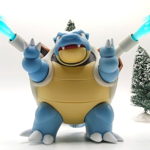 May include: A blue and yellow turtle-like action figure with a brown shell, white claws, and two cannons on its back. The cannons are firing translucent bright blue water blasts. The figure has a wide mouth with visible white teeth.