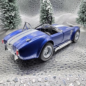 K&ouml;nnte beinhalten: Ein blaues und silbernes Spielzeugautomodell eines klassischen Sportwagens. Das Auto hat einen wei&szlig;en Streifen in der Mitte und einen &Uuml;berrollb&uuml;gel. Das Auto steht auf einem silbernen Hintergrund.