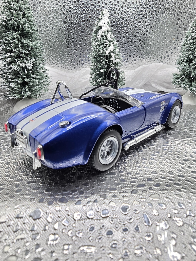 K&ouml;nnte beinhalten: Ein blaues und silbernes Spielzeugautomodell eines klassischen Sportwagens. Das Auto hat einen wei&szlig;en Streifen in der Mitte und einen &Uuml;berrollb&uuml;gel. Das Auto steht auf einem silbernen Hintergrund.