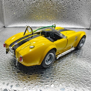 K&ouml;nnte beinhalten: Ein gelbes Spielzeugautomodell eines klassischen Cobra-Sportwagens. Das Auto hat schwarze Rennstreifen und einen detaillierten Innenraum. Das Modell steht auf einem silbernen Hintergrund.