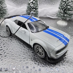 May include: A white toy car with blue racing stripes, featuring black wheels and a black trim. The car is a miniature model, likely an ornament, with a black string for hanging. The background includes miniature snowy trees.