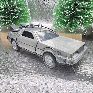 May include: A silver die-cast model of a DeLorean time machine, a car from the Back to the Future movie series. The model has detailed features, including gull-wing doors and a flux capacitor. The car is displayed with miniature Christmas trees.