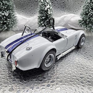 K&ouml;nnte beinhalten: Wei&szlig;es, im Spritzgussverfahren hergestelltes Modell eines klassischen Cobra-Sportwagens mit einem blauen Streifen in der Mitte. Das Auto steht auf einem silbernen Hintergrund.
