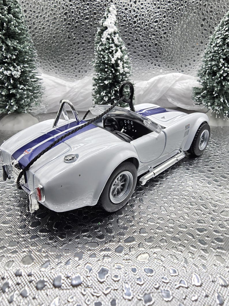 K&ouml;nnte beinhalten: Wei&szlig;es, im Spritzgussverfahren hergestelltes Modell eines klassischen Cobra-Sportwagens mit einem blauen Streifen in der Mitte. Das Auto steht auf einem silbernen Hintergrund.