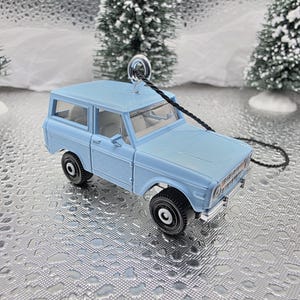 May include: A light blue toy car with black wheels and a silver-colored metal loop for hanging. The car is a miniature replica of a classic SUV. The background includes miniature Christmas trees and a silver surface.