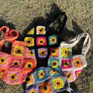 May include: Four crochet tote bags in various colors: orange, black, gray, and off-white. Each bag features a sunflower design within square panels. The bags have matching straps and are displayed on a grassy surface.