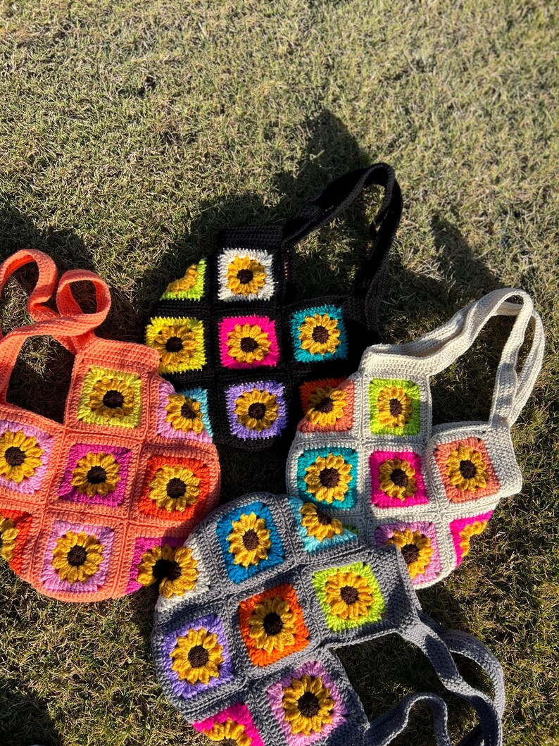 May include: Four crochet tote bags in various colors: orange, black, gray, and off-white. Each bag features a sunflower design within square panels. The bags have matching straps and are displayed on a grassy surface.