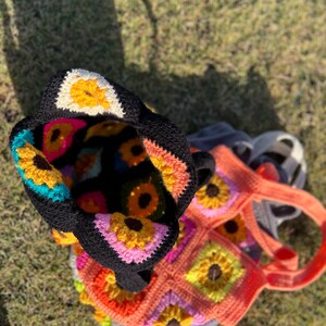 May include: Two crochet tote bags with a sunflower design. One bag is black with colorful sunflowers inside, and the other is orange with a matching sunflower pattern. The bags are handmade and sit on a grassy surface.