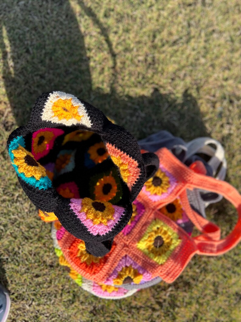 May include: Two crochet tote bags with a sunflower design. One bag is black with colorful sunflowers inside, and the other is orange with a matching sunflower pattern. The bags are handmade and sit on a grassy surface.