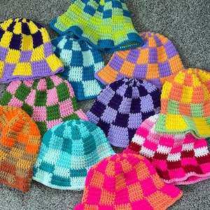 May include: A collection of colorful, handmade crochet bucket hats. Each hat features a checkerboard pattern in various color combinations, including purple and yellow, green and pink, and orange and brown. The hats are displayed on a gray surface.