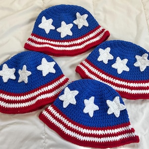 May include: Four blue crochet hats with white stars and red and white striped brims. The hats are arranged on a white surface. Each hat has a different number of white stars.