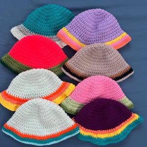 May include: A collection of handmade crochet bucket hats in various colors. The hats feature a classic bucket hat design with a rounded crown and a downward-sloping brim. Colors include teal, red, purple, pink, and white, with striped brims.