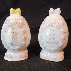 May include: A pair of white ceramic salt and pepper shakers shaped like eggs. The salt shaker features a Precious Moments figurine of a girl holding a lamb. The pepper shaker has the text "The Lord is My Shepherd" on the front. Both shakers have pink and yellow floral designs.