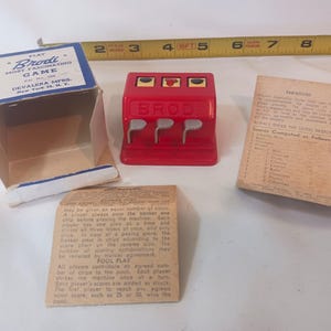 May include: A vintage Brodi game set, featuring a red slot machine-style game with three levers, a cardboard box, and instruction cards. The box is blue and white with the text "Brodi Most Fascinating Game". A yellow measuring tape is in the background.