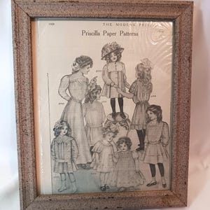 May include: A framed black and white print of a 1909 advertisement for Priscilla Paper Patterns. The ad features seven girls wearing different dresses, with pattern numbers listed next to each girl.