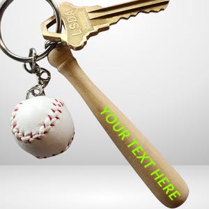 May include: A metal keyring featuring a miniature wooden baseball bat with the neon green text YOUR TEXT HERE printed on the side, a small white baseball with red stitching, and a brass-colored metal key attached to the silver ring.