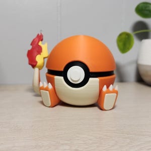 May include: An orange and cream-colored 3D-printed Poke Ball with black accents and white button. The ball has small feet and a flame-like figure attached. A decorative item for fans of the popular franchise.