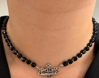 May include: A black beaded necklace with a silver-toned pendant. The pendant is a stylized planet design, encrusted with black gemstones. The necklace is worn around the neck.